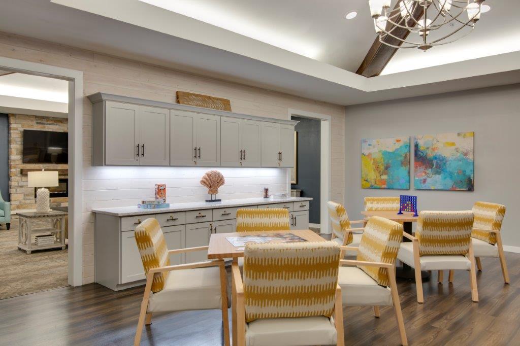 A dinning area with light brown square tables and yellow and orange-striped cushioned chairs around each table, as well as white cupboards along the wall at The Shore at Clear Lake.