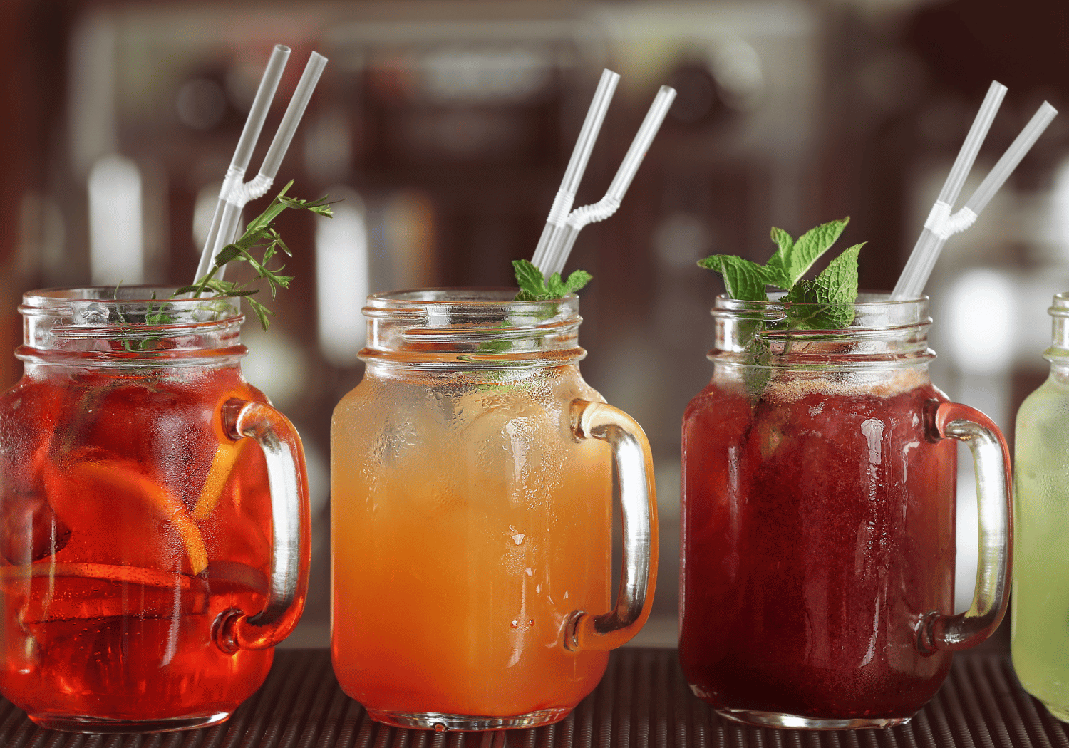 Three mason jar iced drinks with mint garnish and white straws on a bar counter.
