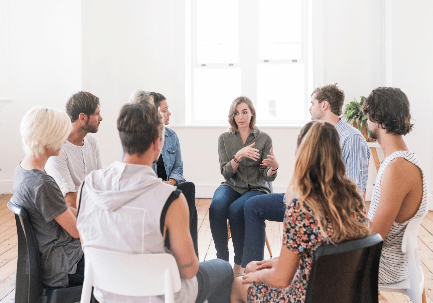Eight adults seated in a circle in a bright room, with a woman in the center speaking and gesturing.