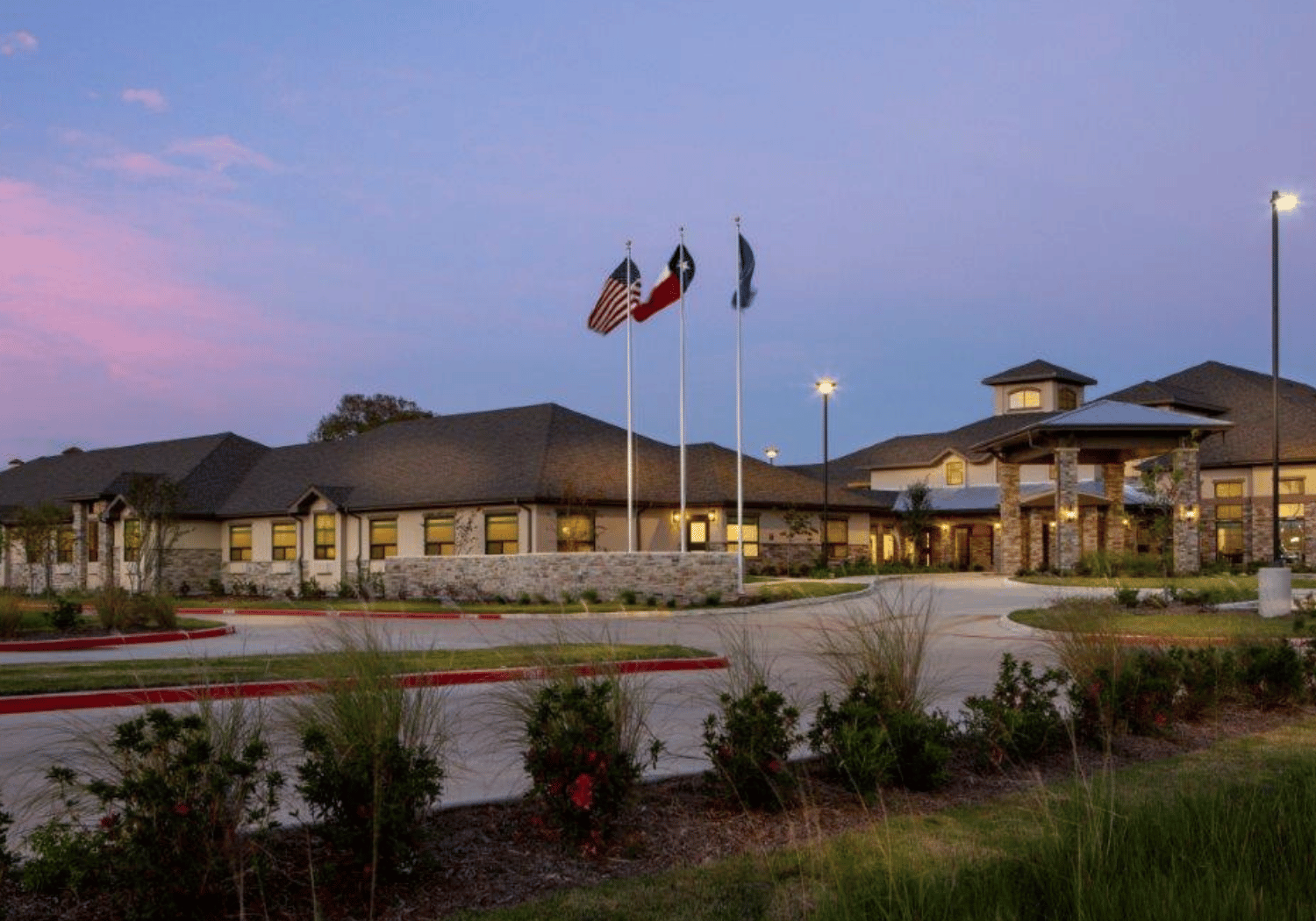 Exterior view of a modern building at dusk with three flagpoles displaying the American, Texas, and organizational flags, surrounded by landscaped grounds.